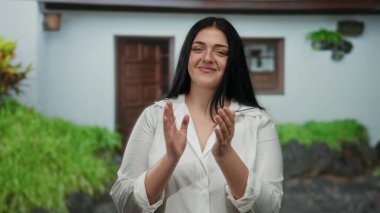 Arka planda bereketli yeşillikle evin dışında el çırpan bir kadın, günlük beyaz gömlekle neşeli bir ifade sergiliyor, huzurlu bir banliyö ortamında güven ve neşe somutlaştırıyor..