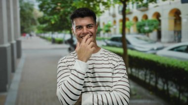 Çizgili tişörtlü genç bir adam şehir caddesinde gülümsüyor, yeşilliklerle çevrili, dışarıda çekici bir şehir portresi sunuyor..