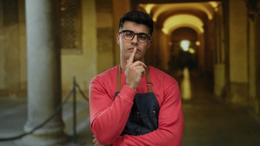 Önlüklü genç adam, tarihi üniversite koridorunda özenle poz veriyor. Mimarinin zarafetini ve geleneksel akademik ortamların çekiciliğini sergiliyor..