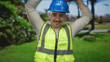 Yansıtıcı yelek giyen ve kasketli genç İspanyol bir adam ellerini başının üstüne koyup kalp hareketi yapıyor..