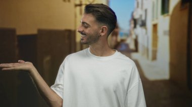 Beyaz tişörtlü ve neşeli gülümsemeli bir adam güneşli eski kasaba caddesinde avuçlarını ileri açık tutar; sıcak karşılama.