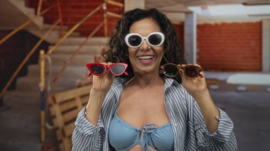 İnşaat alanında elinde güneş gözlüğü tutan orta yaşlı İspanyol kadın. Rahat çizgili gömleği ve canlı atmosferiyle güvenle gülümsüyor..