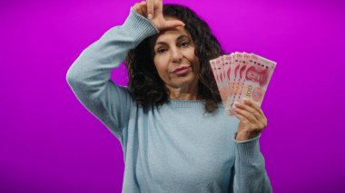 Bir kadın Çin Yuan banknotlarını tutarken ezik jestler yapıyor, izole edilmiş mor duvarlara karşı duruyor, duygularını ve finansal kavramlarını ifade ediyor..