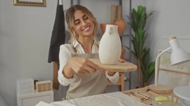 Önlük takan sarışın kadın, çömlek atölyesindeki ahşap döner tablonun üzerinde kilden vazo tutuyor..