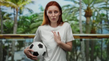 Parkta kızıl saçlı bir kadın elinde bir futbol topu tutuyor, kafası karışmış ve beyaz bir gömlek giyiyor, canlı yeşillikle çevrili..
