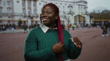 Pembe örgülü bir kadın, tarihi bir bina ve arka planda hareketli bir kalabalıkla güvenli bir şekilde gülümseyerek, canlı bir şehir atmosferini yakalıyor..