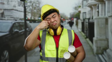 Güvenlik yeleği ve miğferi olan Asyalı genç adam komik bir şekilde şehir sokaklarında hareket ediyor, elinde neşeli bir ifadeyle planlar tutuyor..