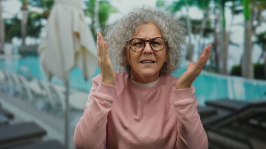 Bir kadın havuz kenarında, pembe gömlekli, gri bukleli ve gözlüklü, tropikal tatil seti sergiliyor..