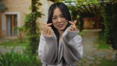 Serin bahçede gülümseyen bir kadın yemyeşil ve taş patikalı, rahat bir kazak giyen ve elleriyle kalp hareketi yapan, sıcaklığı ve mutluluğu ileten bir kadın..