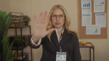 Ofiste duran, gözlük takan ve arkaplan çizelgesi olan siyah takım elbiseli bir kadın, kapalı alanda profesyonel bir ortam izlenimi veriyor..