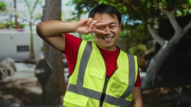Dışarıda güneşli bir günde parkta gülümseyen güvenlik yeleği giyen genç Çinli bir adam.