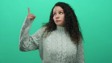 Süveter giyen genç İspanyol kadın, yeşil duvar arkaplanını işaret ediyor. Onaylamadığını belirtiyor ve yön gösteriyor..