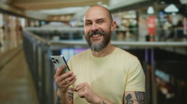 Sakallı bir adam modern bir alışveriş merkezinde akıllı telefon kullanıyor. Şehir yaşamını geniş ışıklandırmalı, hareketli bir ev ortamında sergiliyor..