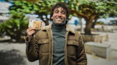 Ağaç ve kentsel geçmişe sahip bir şehir caddesinde elinde 50 avroluk banknot tutan gülümseyen İspanyol adam, açık havada canlı bir şekilde günlük kıyafetlerini ve finansal başarılarını sergiliyor..