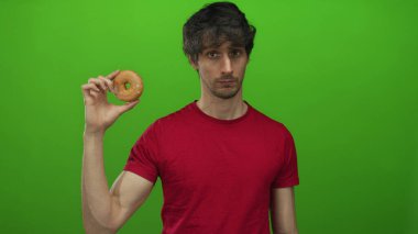 Adam parlak yeşil stüdyoda kameraya jöleli donut tutar tarafsız ifade ile; açlık arzu iştah arzusu.