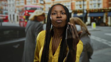 Şehir caddesinde sarı tişörtlü bir kadın, kırmızı otobüsü ve arka planda binaları var. Şehir ortamında örülmüş saçları olan, modern şehir yaşam tarzını gösteren ciddi görünümlü bir kadın.