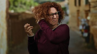 Şehir ortamında kıvırcık saçlarını düzelten bir kadın, gözlük takarken hoşnutsuzluğunu dile getiriyor, bir şehir caddesinde yakalandı..