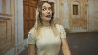 Eski bir üniversitenin önünde duran bir kadın, el ele tutuşarak şaşkınlığını dile getiriyor, etrafı tarihi mimariyle çevrili, parlak gün ışığında akademik bir ortama işaret ediyor..