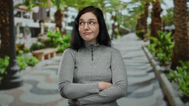 Açık havada gülümseyen esmer saçlı bir kadın ve palmiye çizgili bir sokakta gri bir süveter giyen yeşil bitkilerle canlı bir şehir zemini oluşturan bir kadın..