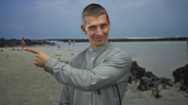 Baş parmaklı ve işaret eden bir adam güneşli bir sahilde gülümsüyor. Arka planda açık deniz ve kayalar var. Olumlu ve rahat bir açık hava yaratıyor..