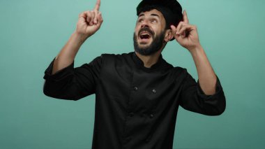 Siyah üniformalı İspanyol erkek aşçı, yeşil bir arka plana doğru bakıyor, coşku ve mutfak uzmanlığını ifade ediyor, neşeyle gülümsüyor..