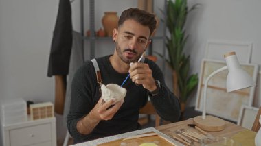 Stüdyodaki ahşap masanın üzerine fırçayla boyarken elinde kilden bir kase tutan adam; yaratıcı odak.