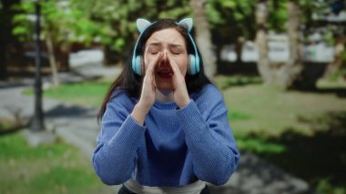 Kulaklık takan ve yeşil park ortamında özellikle el kol hareketi yapan bir kadın dışarıda duygusal bir ifade sergiliyor..