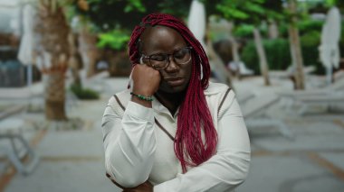 Bir kadın şehir caddesinde elleri çenesinde dikiliyor düşünceli görünüyor, gözlükler takıyor ve örgü örüyor, ağaçlar ve şehir manzarasıyla çevrili..