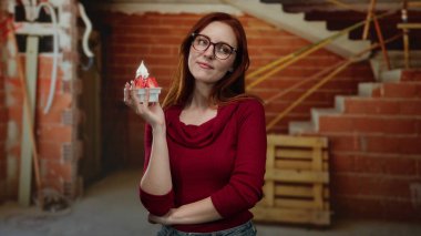 İnşaat alanında elinde dondurma tutan kırmızı tuğlalı arka plan, kırmızı süveter ve gözlük giyen, neşeli bir ifade sergileyen, yaşam tarzı konsepti için kapalı alan ayarlı bir kadın..