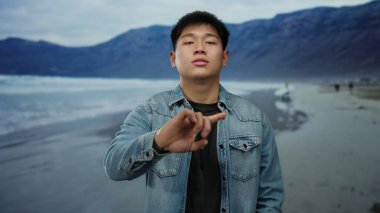 Sahilde genç bir adam kot ceketli parmaklarıyla okyanusa ve arka planda dağlara doğru hareket ediyor.