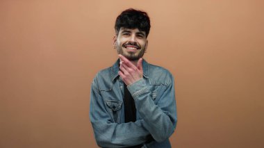 Kot ceketli genç adam, düz kahverengi bir arka plan önünde kendinden emin bir şekilde gülümsüyor..