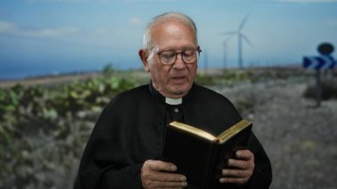 Arka planda yel değirmenleri olan bir açık alanda kitap tutan kıdemli bir rahip, dinsel bir açık hava manzarasını ve huzurlu bir kırsal ortamı gösteriyor..