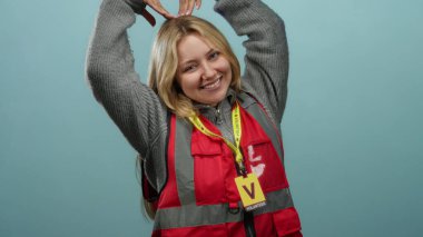 Kırmızı gönüllü yelek giymiş, gülümseyen sarışın kadın, elleri mavi duvar arkasında kalp sembolü yapıyor..
