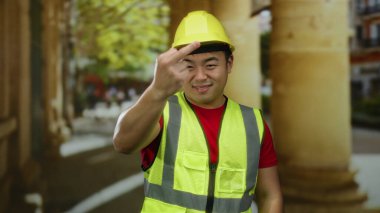 Kasketli ve yansıtıcı yelek giyen adam arka planda sütunlar ve ağaçlarla şehir caddesinde jest yapıyor..