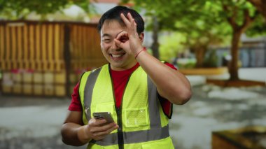 Parkta gülümseyen bir adam açık havada sarı yelek giyip akıllı telefondan mesajlaşırken iletişim ve aktiviteyi temsil ediyor..