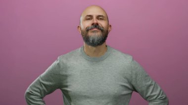 Kel kafalı sakallı adam güvenli bir şekilde pembe arka plana karşı duruyor. Pozitif ve ulaşılabilir bir hava yayıyor.