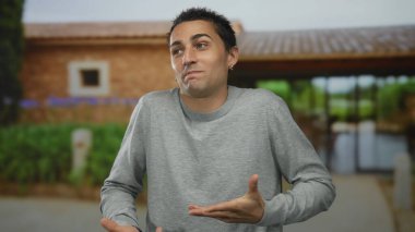Kırsal bir binanın önünde, gri bir süveter ve küpe takmış gevşek bir ifadeyle el kol hareketi yapan genç bir İspanyol açık hava havasını yakalıyor..