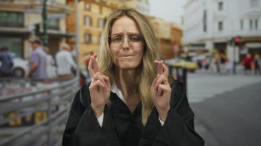 Sarı saçlı ve gözlüklü bir kadın yargıç şehir sokaklarında parmaklarını çapraz yapıp siyah bir üniforma giyip şehir içinde kararlılığını ve odak noktasını gösteriyor..