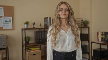 Genç sarışın bir kadın organize bir ofiste raflar, dosyalar ve bitkilerle profesyonel bir iş ortamı yaratıyor.