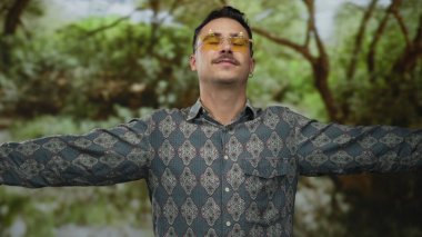 Genç İspanyol adam desenli gömlek ve güneş gözlüğü takıyor. Yeşillik yeşil bir parkta kollarını açmış, açık havada özgürlük ve neşe hissi yayıyor..