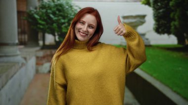 Sarı kazaklı, kızıl saçlı kadın şehir caddesinde, açık havada canlı, yemyeşil bir sahne sergiliyor..