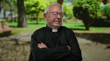 Siyah rahip kollarını kavuşturmuş, güneşli bir günde yeşil bir parkta gülümsüyor..