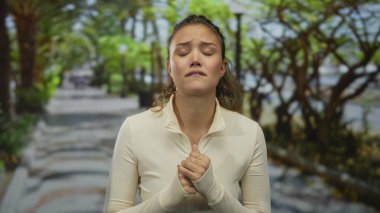 Parktaki İspanyol kadın duygusal görünüyor. Arka planda yeşil yapraklar var. Açık renk ceket giyiyor. Üzüntüsünü dile getiriyor..