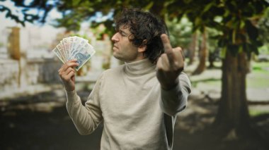 Orta parmağını gösteren İspanyol bir adam parkın dışında zloti banknotları tutarken, arka planda ağaçlara olan öfkesini gün ışığında dile getiriyor..