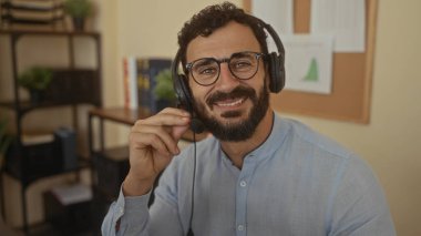 Ofis ortamında kulaklık takarken gülümseyen sakallı İspanyol bir adam profesyonel bir iş ortamı öneriyor..