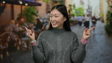 Şehirli bir terasta gülümseyen ve parmakla işaret eden bir kadın hareketli bir şehir sokak ortamında gri bir süveter giyip açık hava ortamı sergiliyor..