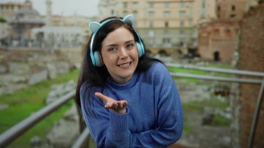 Kulaklıklı esmer kadın, antik Roma harabelerinin arasında gülümsüyor, güzel ve dingin bir şekilde tarihi bir sahne sergiliyor..