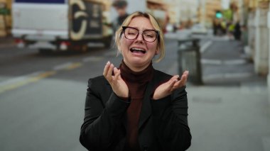 Arka planda bulanık yayalar ve şehir araçlarıyla çevrili, gözlüklü ve koyu renk ceketli, hareketli bir caddede duygusal olarak ağlayan bir kadın..