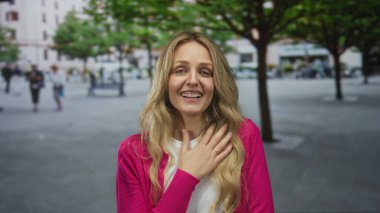 Açık havada gülümseyen genç sarışın kadın canlı pembe bir süveter ve uzun saçlı, ağaçlarla çevrili ve arka planda bulanık şehir hayatı, mutluluk saçan.