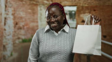 Bir inşaat alanında gülümseyen ve beyaz bir alışveriş torbasını inceleyen bir kadın, kırsal tuğla bir arka planda dururken gözlüğünü ve gri kazağını sergiliyor..